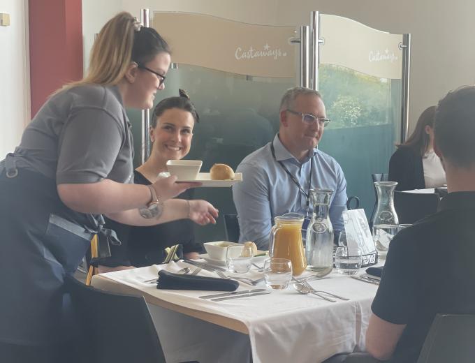 A student from Ganton School smiling and serving food to a table of guests.