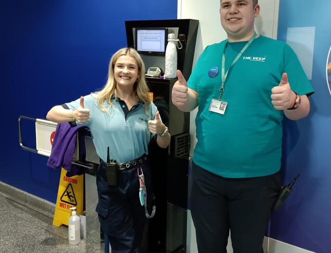 Work experience student, Jacob, stood smiling and both thumbs up with one of The Deep's Guides, Becki, also stood smiling with both thumbs up. They are stood in front of the entrance turnstile.