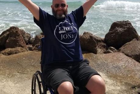 Jon Simpson on a beach in his wheelchair with happy raised arms