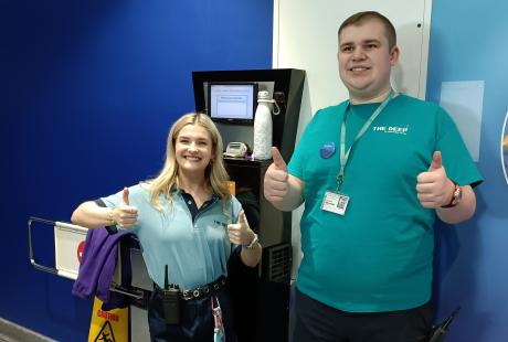 Work experience student, Jacob, stood smiling and both thumbs up with one of The Deep's Guides, Becki, also stood smiling with both thumbs up. They are stood in front of the entrance turnstile.